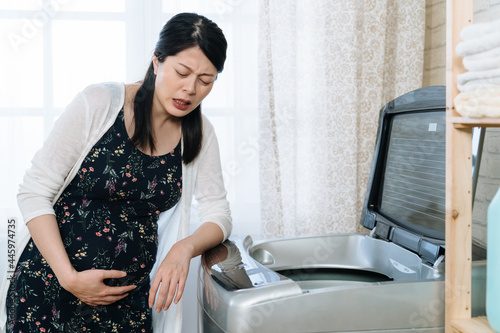 Young asian chinese pregnant woman suffering from abdominal pain at home laundry room. illness maternity housewife having stomach ache doing housework time. sick future mom by washing machine
