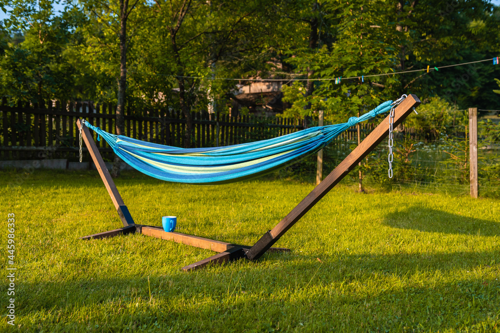 Blue, wooden hammock and blue cup standing on green grass in a rural scenery in the evening sun