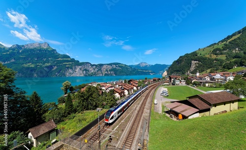 Summer scenery of beautiful Lake Lucerne on a sunny day, with a train traveling on the railway thru Sisikon Village by Lake Uri & majestic mountains in background under blue clear sky in Switzerland photo