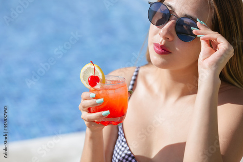 young woman in bikini and sunglasses holding refreshing drink by pool on sunny day