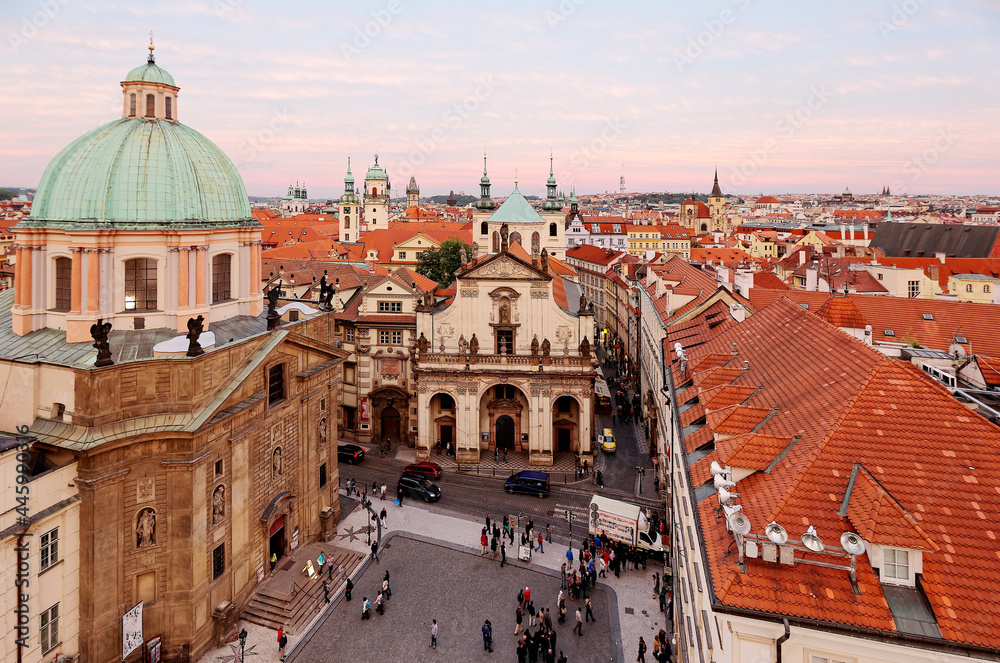 Panoramic view from the Bridge Tower overlooking a square in Prague Old Town, Czech, with a skyline of majestic domes, red tiled roofs and towering spires of historic buildings under rosy sunset sky