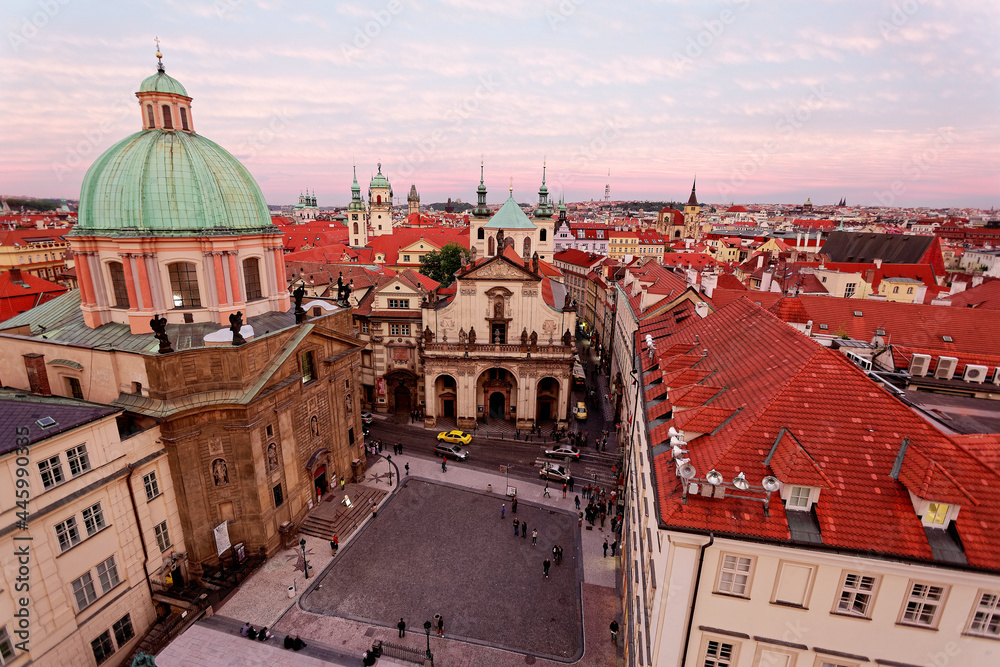 Panoramic view from the Bridge Tower overlooking a square in Prague Old Town, Czech, with a skyline of majestic domes, red tiled roofs and towering spires of historic buildings under rosy sunset sky