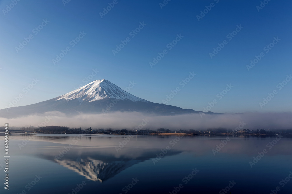 Beautiful scenery of snow capped Mount Fuji reflected in the peaceful lake water under blue clear sky with low stratus clouds at the foothill on a sunny winter morning at Kawaguchiko, Yamanashi, Japan
