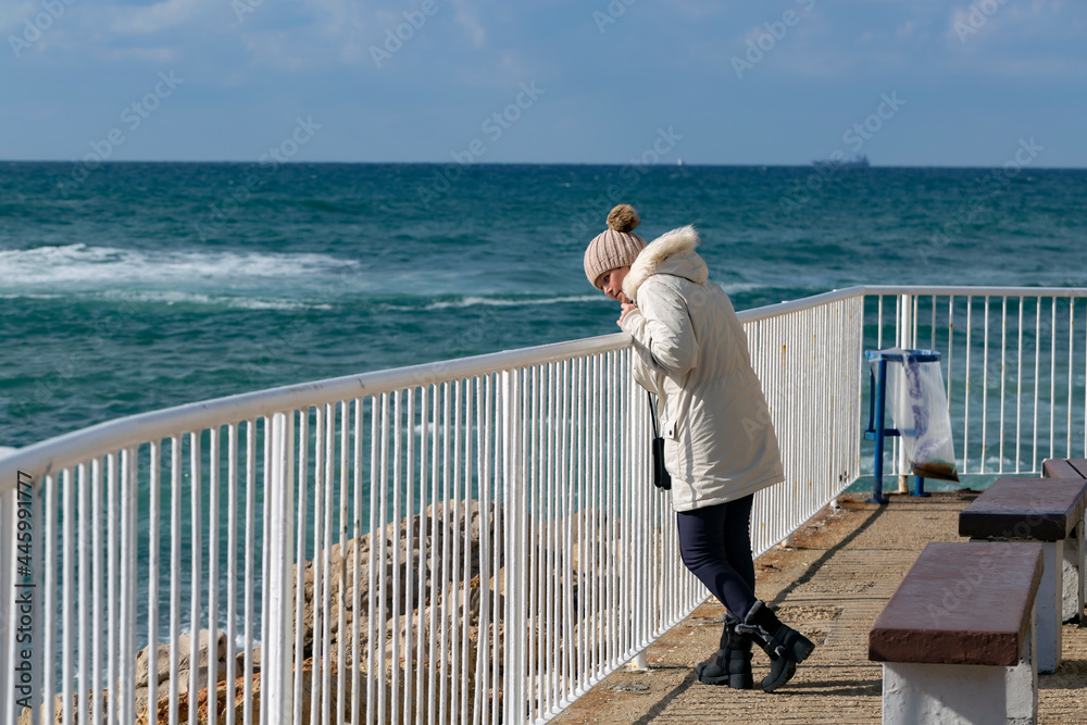 Happy caucasian american woman standing on the beach in a white down jacket and knitting hat with a view of the blue sea and enjoying freedom. Emotional woman walking on white pier against the sea