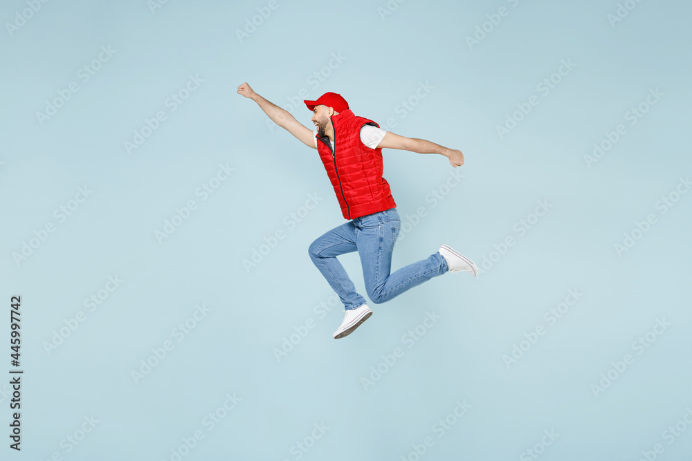 Full size body length side view delivery guy employee man in red cap white T-shirt vest uniform work dealer courier jump run go walk isolated on pastel blue color background studio. Service concept