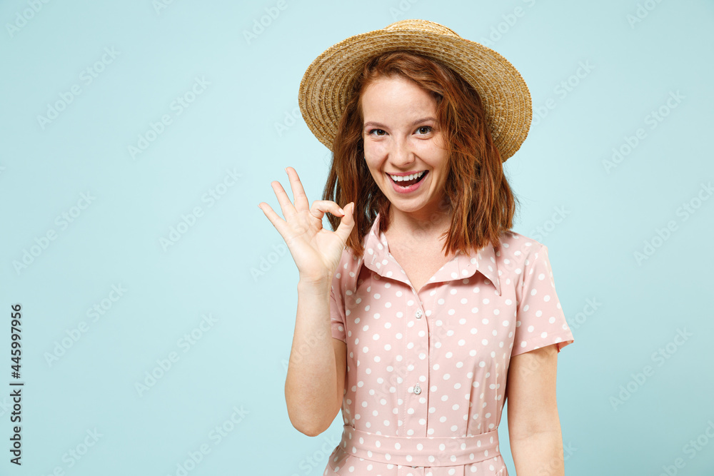 Satisfied smiling young redhead curly woman 20s show ok okay wears casual pink dress straw hat look camera isolated on pastel blue color background studio portrait. People emotions lifestyle concept.