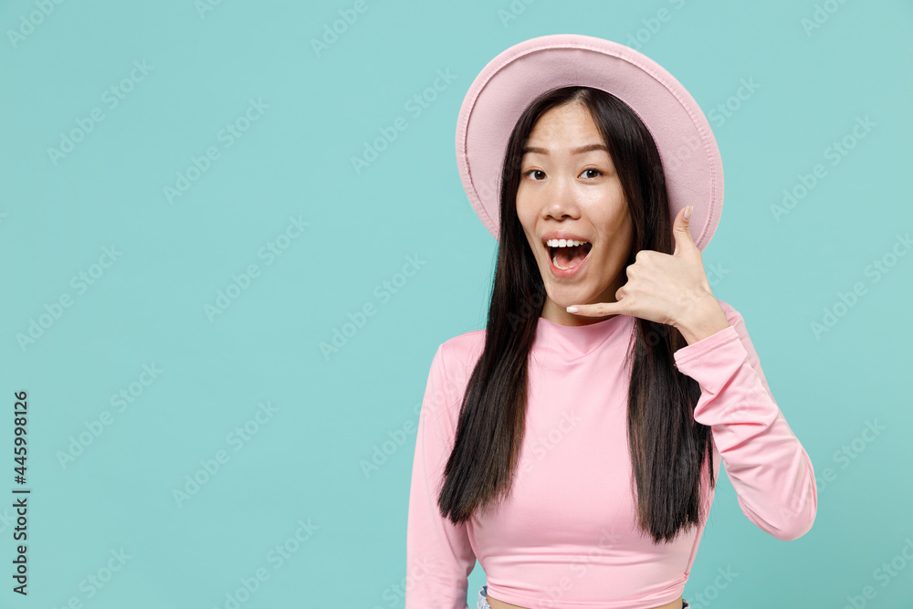 Smiling happy cheery young brunette asian woman 20s wears pink clothes hat doing phone gesture like says call me back isolated on pastel blue color background studio portrait. People emotions concept Smiling happy cheery young brunette asian woman 20s wears pink clothes hat doing phone gesture like says call me back isolated on pastel blue color background studio portrait. People emotions concept
