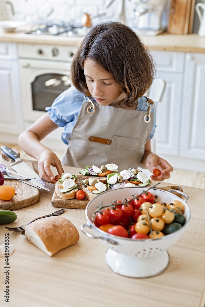 Cute brunette teen makes sandwiches in the domestic kitchen. Beautiful girl is cooking in the kitchen. Attractive girl in apron on a light color home kitchen background. One person. Domestic life Cute brunette teen makes sandwiches in the domestic kitchen. Beautiful girl is cooking in the kitchen. Attractive girl in apron on a light color home kitchen background. One person. Domestic life