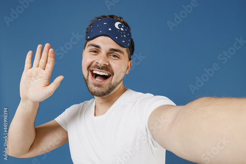 Close up friendly young man 20s in pajamas jam sleep mask rest relax at home doing selfie shot on mobile phone waving hand say hello isolated on dark blue background. Good mood night bedtime concept.