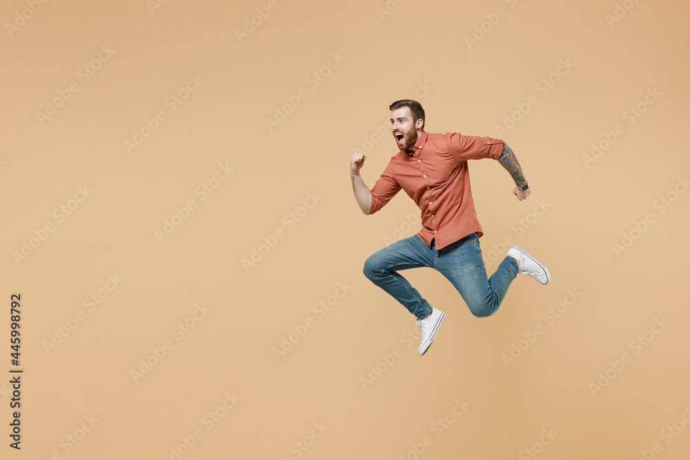 Full size body length handsome fun tatooed young brunet man 20s short haircut open mouth wears apricot shirt jump like running hurry up look aside isolated on pastel orange background studio portrait