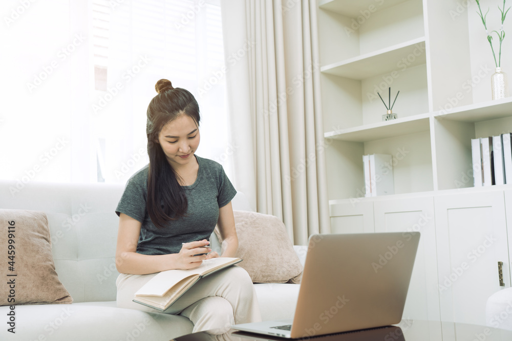 Asian woman sits on the sofa while taking notes from her computer screen while working or studying online at home. Asian woman sits on the sofa while taking notes from her computer screen while working or studying online at home.