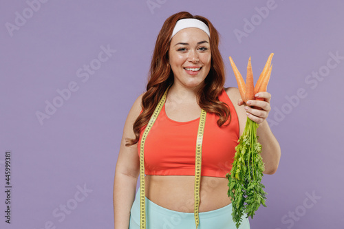 Young vegeterian chubby overweight plus size big fat fit woman in red top measuring tape warm up training hold raw organic carrot bunch isolated on purple background home gym Workout sport concept