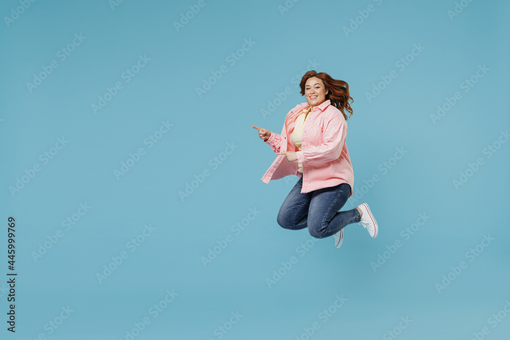 Full length young redhead chubby overweight woman 30s wearing in pink shirt jeans jumping high point index finger aside on workspace advertisement free place isolated on pastel blue background studio. Full length young redhead chubby overweight woman 30s wearing in pink shirt jeans jumping high point index finger aside on workspace advertisement free place isolated on pastel blue background studio.