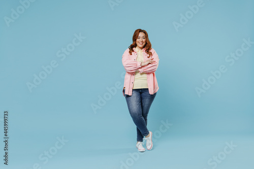 Full length young fun smiling redhead chubby overweight woman 30s with curly hair wear in pink shirt jeans casual clothes hold hands crossed folded isolated on pastel blue background studio portrait