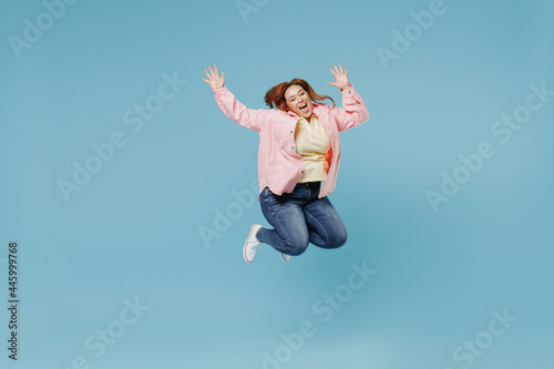 Full length young smiling redhead chubby overweight woman 30s with curly hair wear in pink shirt jeans casual clothes do winner gesture clench fist isolated on pastel blue background studio portrait