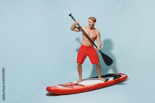 Sticker Full length young sexy strong sporty blond man 20s wear red shorts swimsuit standing sup board paddle surfing look camera isolated on pastel blue background