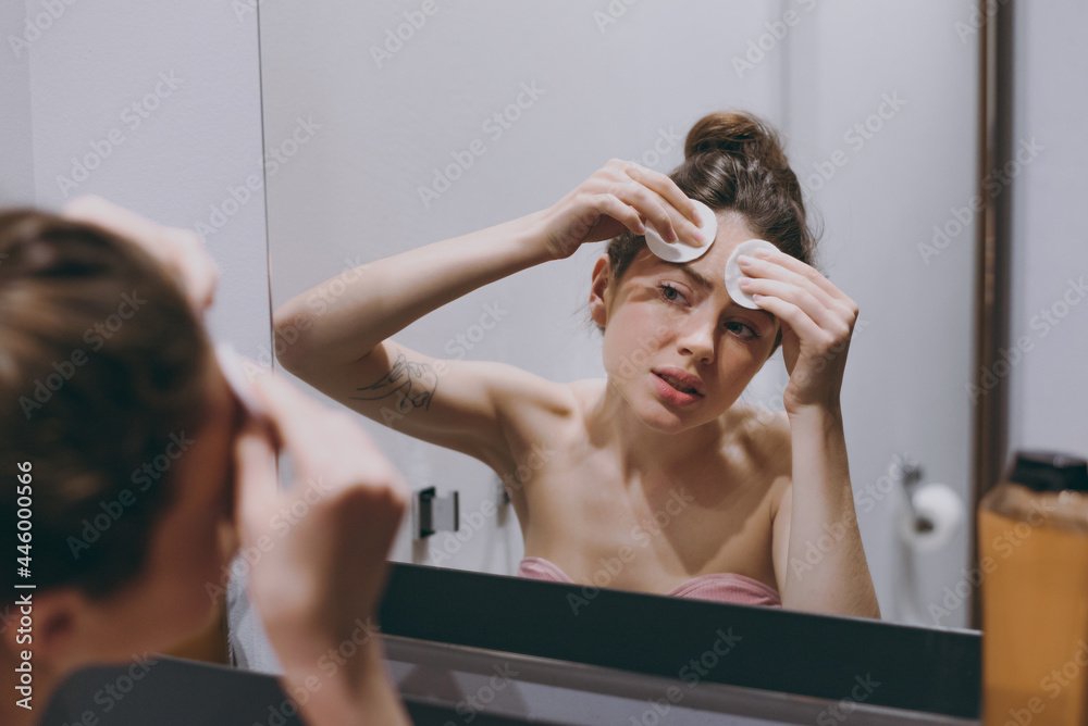 Naklejka premium Disappointed sad half naked young woman 20s wearing bra wipes face with cotton swabs tonic lotion reflected in mirror in bathroom do morning routine Skin care healthcare cosmetic procedures concept..