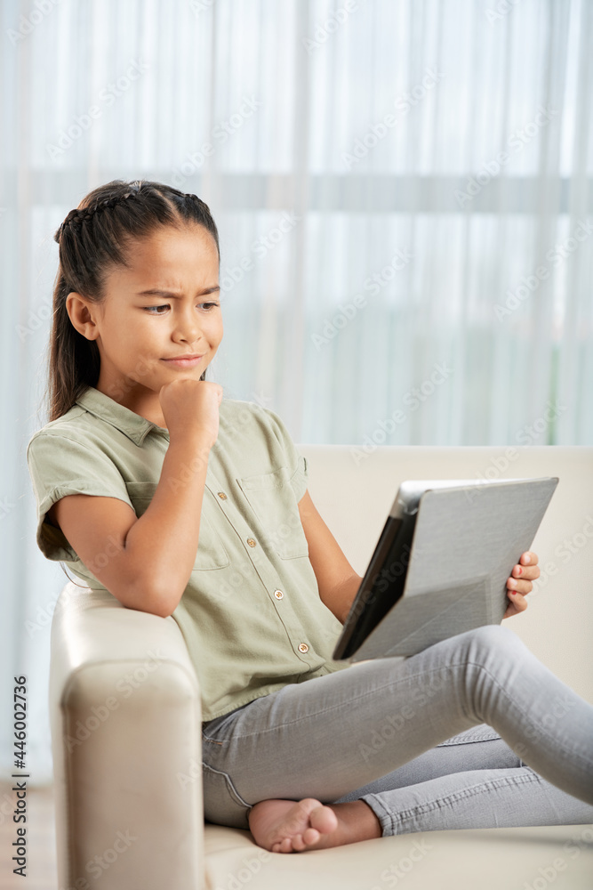 Little girl sitting on sofa with pensive sight and looking at digital tablet, she studying online at home Little girl sitting on sofa with pensive sight and looking at digital tablet, she studying online at home