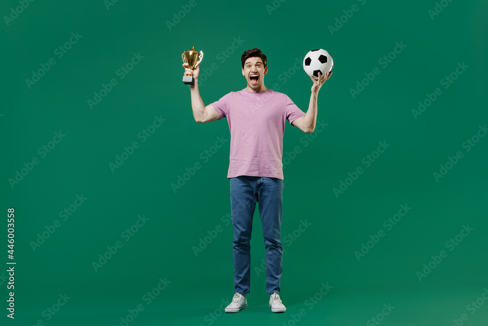 Full size body length young fun man fan wears pink t-shirt cheer up support football sport team hold in hand soccer ball cup watch tv live stream scream isolated on dark green color background studio.