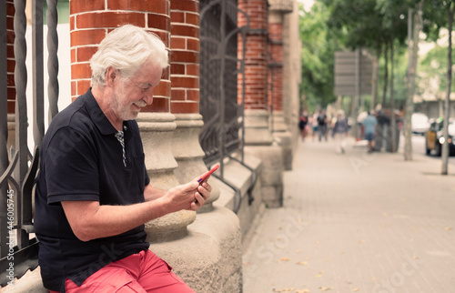 Senior man sitting outdoors in city street using mobile phone, elderly retired smiles