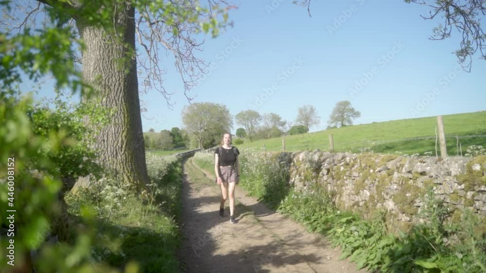 Slow-mo of blonde woman smiling walking towards camera along dirt track Ashbourne, Peak District, England. Blue, clear skies, green fields, trees and overgrown hedgerows. Slow-mo of blonde woman smiling walking towards camera along dirt track Ashbourne, Peak District, England. Blue, clear skies, green fields, trees and overgrown hedgerows.