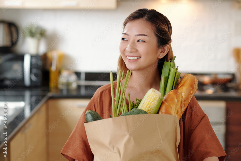 Portrait of happy pretty young woman standing in kitchen with paper bag of fresh groceries like bread and vegetables Portrait of happy pretty young woman standing in kitchen with paper bag of fresh groceries like bread and vegetables