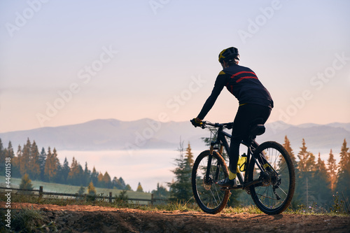 Silhouette of cyclist riding bike with coniferous trees and hills on background. Back view of man bicyclist enjoying bicycle ride in mountains in the morning. Concept of biking and active leisure.