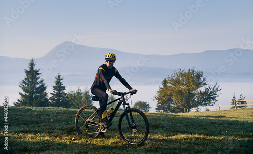 Smiling man cyclist in cycling suit riding bicycle on grassy hill. Male bicyclist in safety helmet enjoying the view of majestic mountains during bicycle ride. Concept of sport, bicycling and nature.