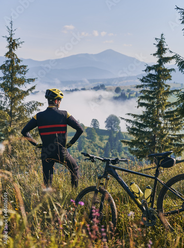 Fototapeta Naklejka Na Ścianę i Meble -  Back view on a cyclist wearing uniform and helmet, standing in grass near mountain bike, watching beautiful mountains landscape. Early morning in foggy sunny mountains. Concept of unity with nature