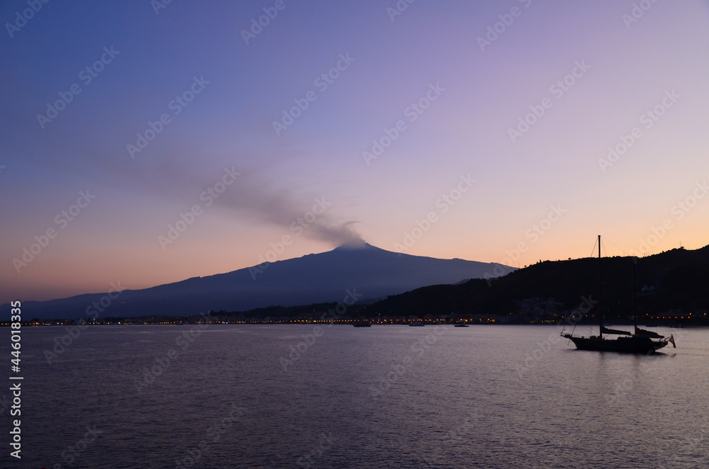 A sunset view of Etna volcano mountain and a boat silhouette on the sea.