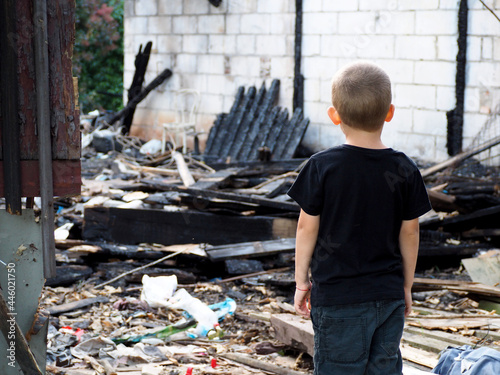 a little boy in a black T-shirt and pants stands with his back against the background of a burned-out house