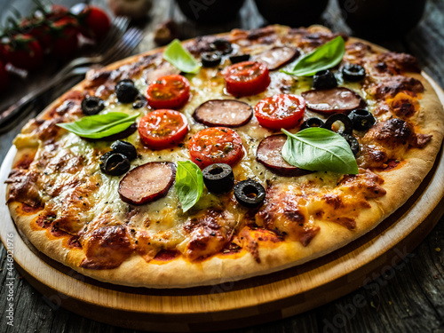 Pizza with white mushrooms, sausage, tomatoes, black olives, parmesan and mozzarella on wooden background 