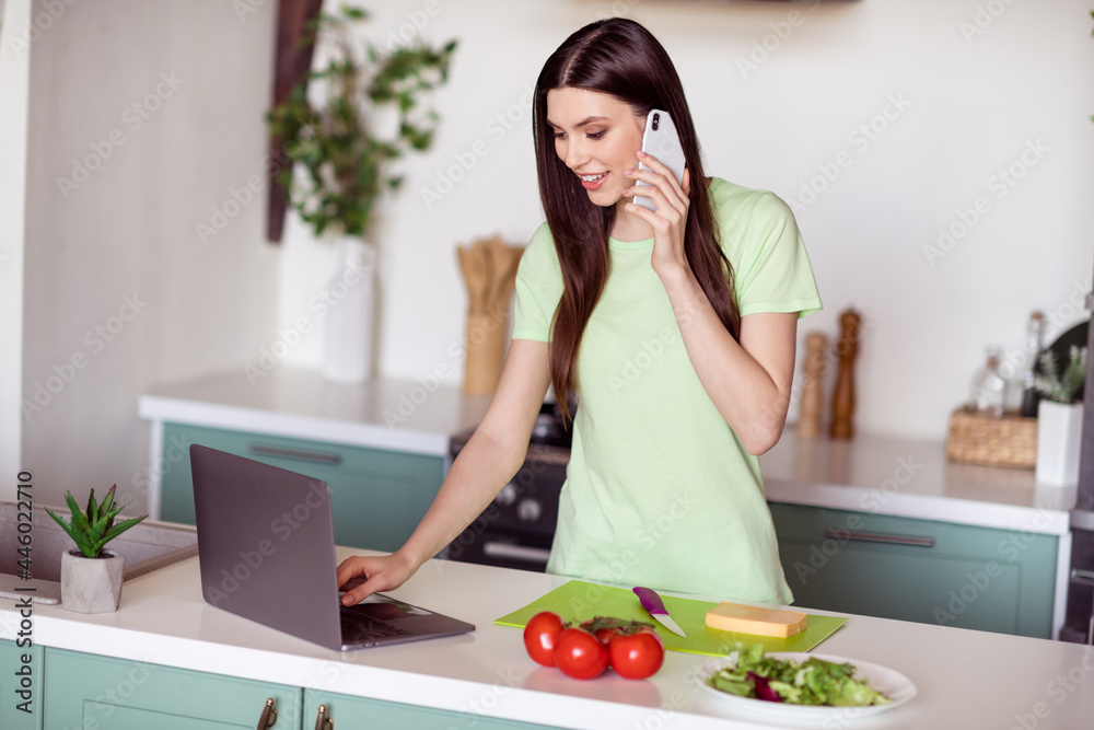 Photo of adorable busy young woman dressed green t-shirt cooking salad talking typing modern gadgets indoors house home room