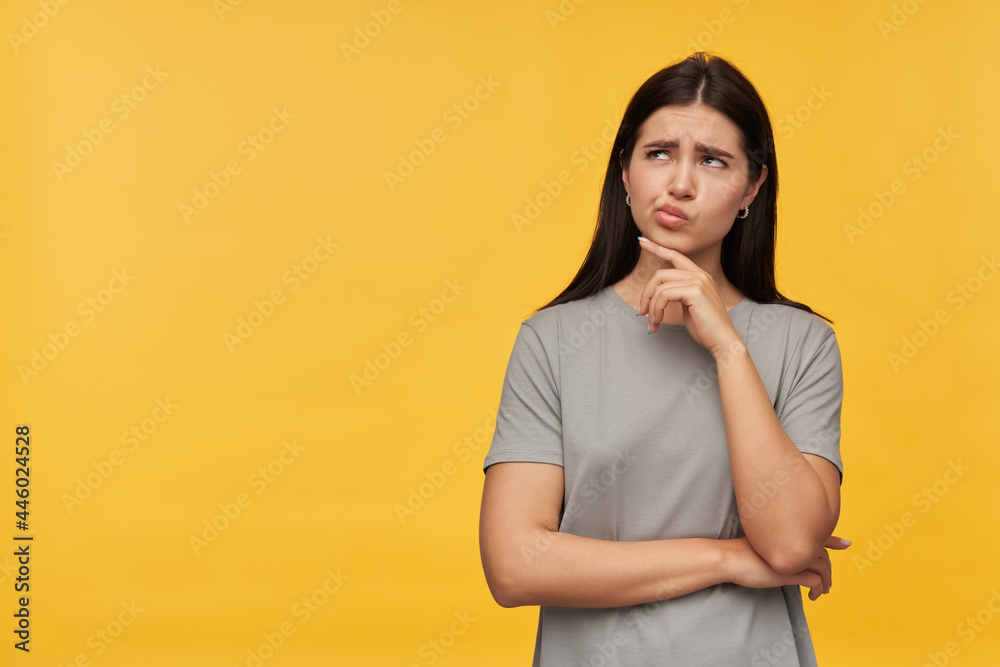 Thoughtful pretty young woman with dark hair in gray tshirt touching her chin and looking away to the side at empty space over yellow background Thoughtful pretty young woman with dark hair in gray tshirt touching her chin and looking away to the side at empty space over yellow background