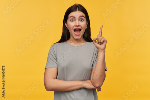 Amazed excited young woman with dark hair and opened mouth in gray tshirt pointing up by finger and having an idea over yellow background