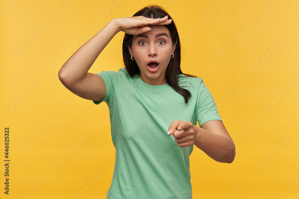 Surprised shocked brunette young woman in mint tshirt keeps hand on forehead looking far away and pointing on you at camera over yellow background Surprised shocked brunette young woman in mint tshirt keeps hand on forehead looking far away and pointing on you at camera over yellow background