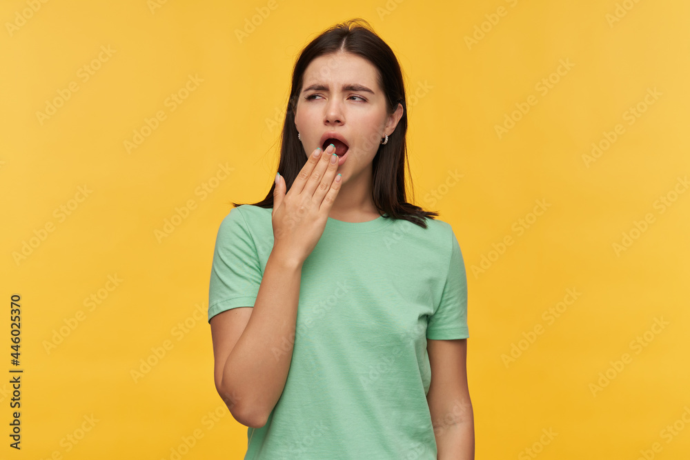Sleepy exhausted brunette young woman in mint tshirt covered mouth by hand and yawning isolated over yellow background Sleepy exhausted brunette young woman in mint tshirt covered mouth by hand and yawning isolated over yellow background