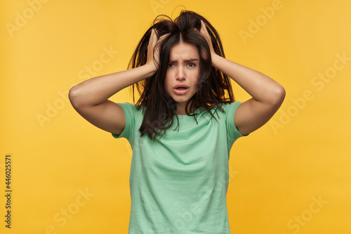 Sleepy pretty young woman with dark messy hair in mint tshirt keeps hands on head and feels oversleep isolated over yellow background