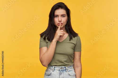 Happy beautiful brunette young woman in casual clothes keeps fingers on lips and showing silence gesture over yellow background Looking at camera