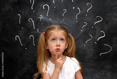Blond schoolgirl girl on a black background of chalk board with a questioning expression on her face. Question marks written in chalk on a blackboard.