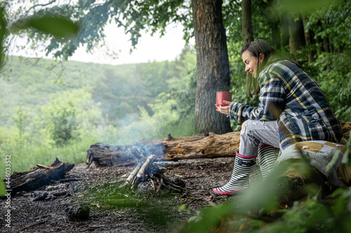 An attractive girl with a cup in her hand warms up near a fire in the forest.