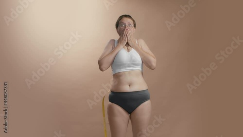 Full figured girl in white bra and black briefs with measure tape to know the volume of her waist. Surprise face expression. Beige background slow motion high quality video.