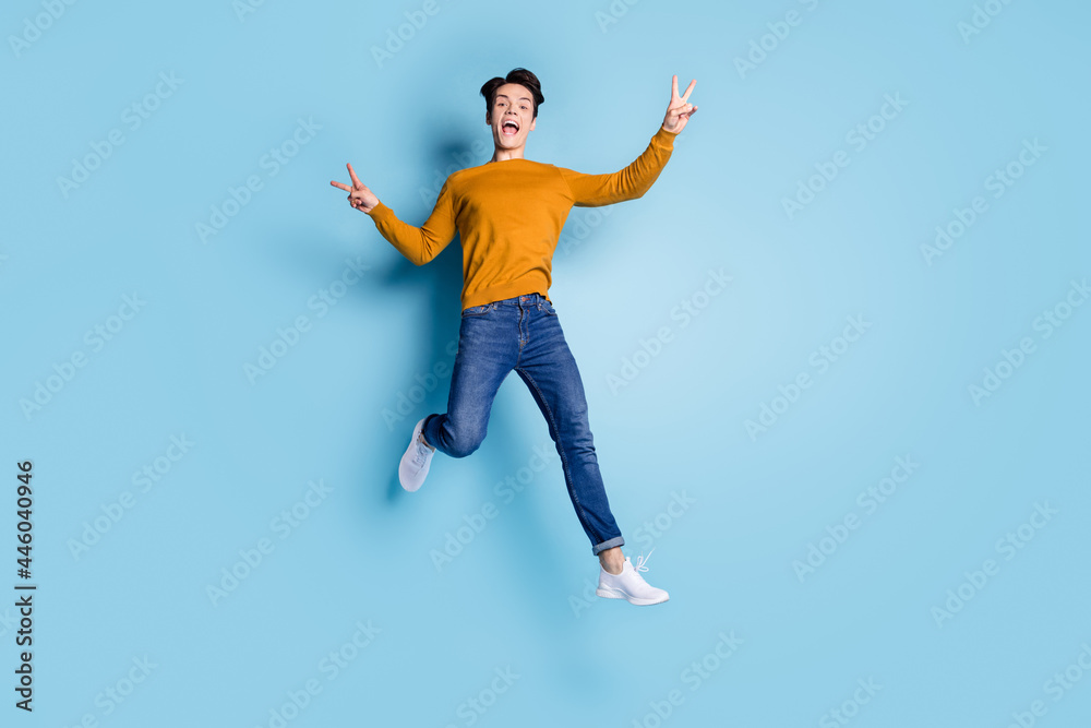 Full size photo of crazy brunet millennial guy jump show v-sign wear sweater jeans isolated on blue color background Full size photo of crazy brunet millennial guy jump show v-sign wear sweater jeans isolated on blue color background