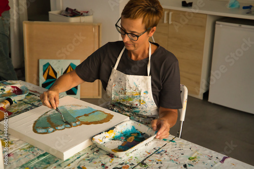 Mature woman with glasses sitting painting a picture of a butterfly in her painting studio.