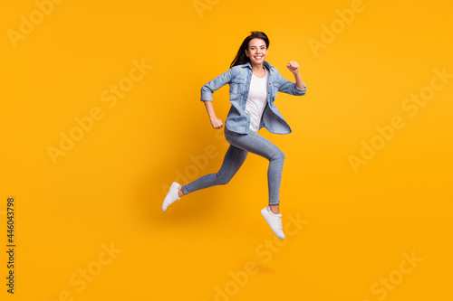Full size profile photo of cool optimistic brunette lady run jump wear blue shirt jeans sneakers isolated on yellow background