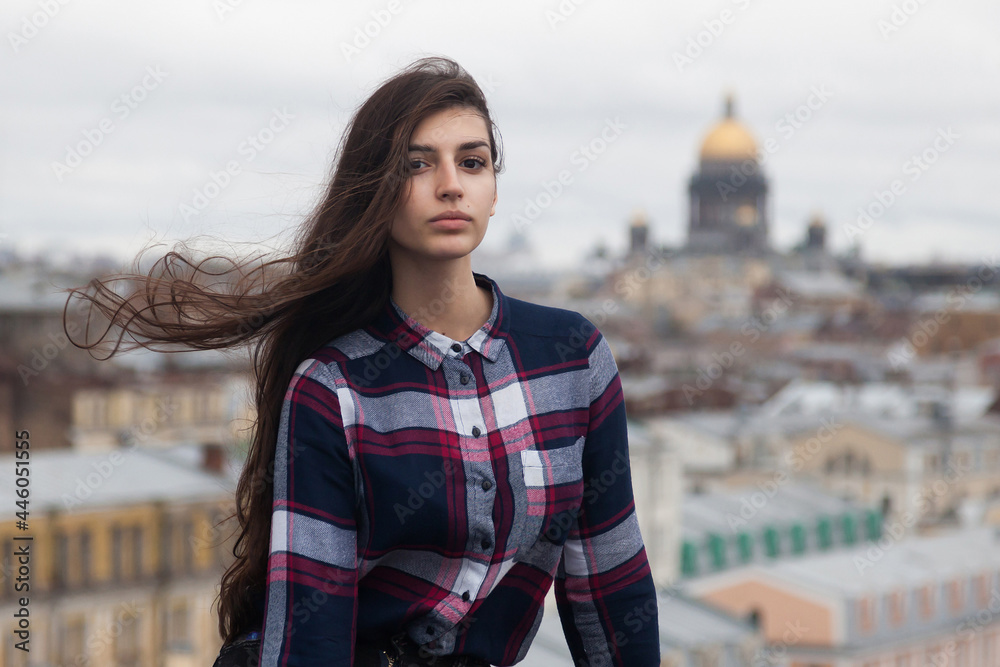 portrait of an Armenian girl with fluttering long black hair in a checkered shirt and jeans on a rooftop in the center of St. Petersburg