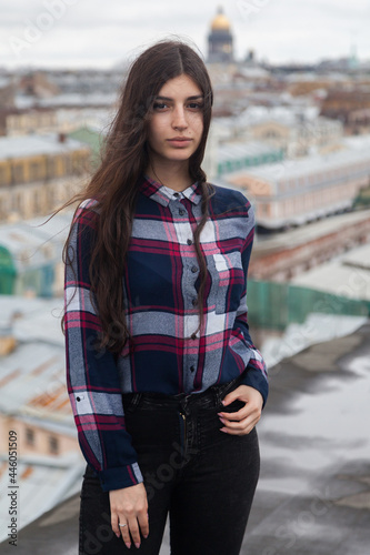 An Armenian girl with long black hair in a plaid shirt stands on a rooftop in the center of St. Petersburg