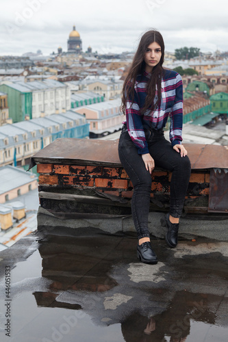 An Armenian girl with long black hair in a plaid shirt stands on a rooftop in the center of St. Petersburg