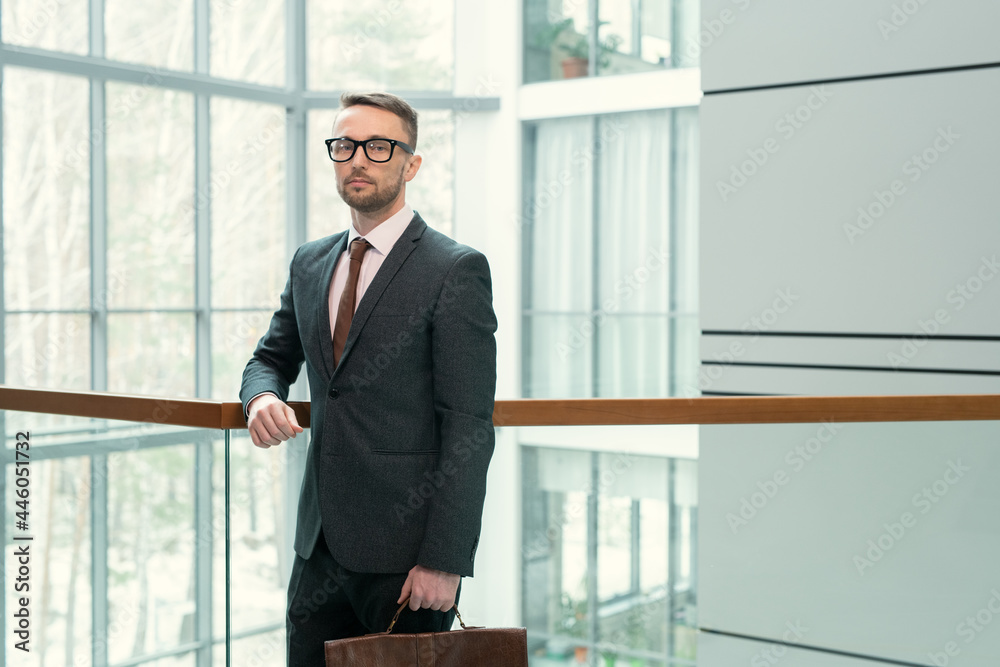 Portrait of elegant businessman in suit looking at camera while standing at office corridor with glass walls