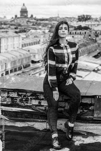 An Armenian girl with flowing long black hair in a checkered shirt sits on a rooftop in the center of St. Petersburg